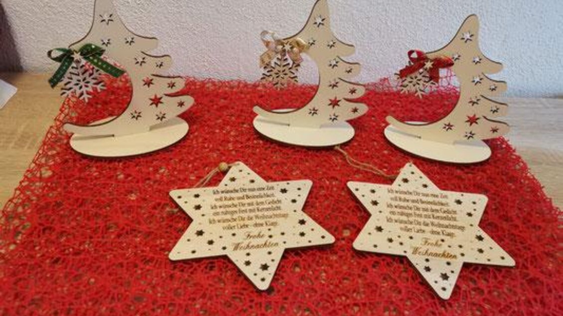 Holzfiguren in Form von Weihnachtsbäumen und Sternen liegen auf einer roten Unterlage. Die Figuren sind mit bunten Schleifen und Schriftzügen verziert. | © KM Lasergravuren