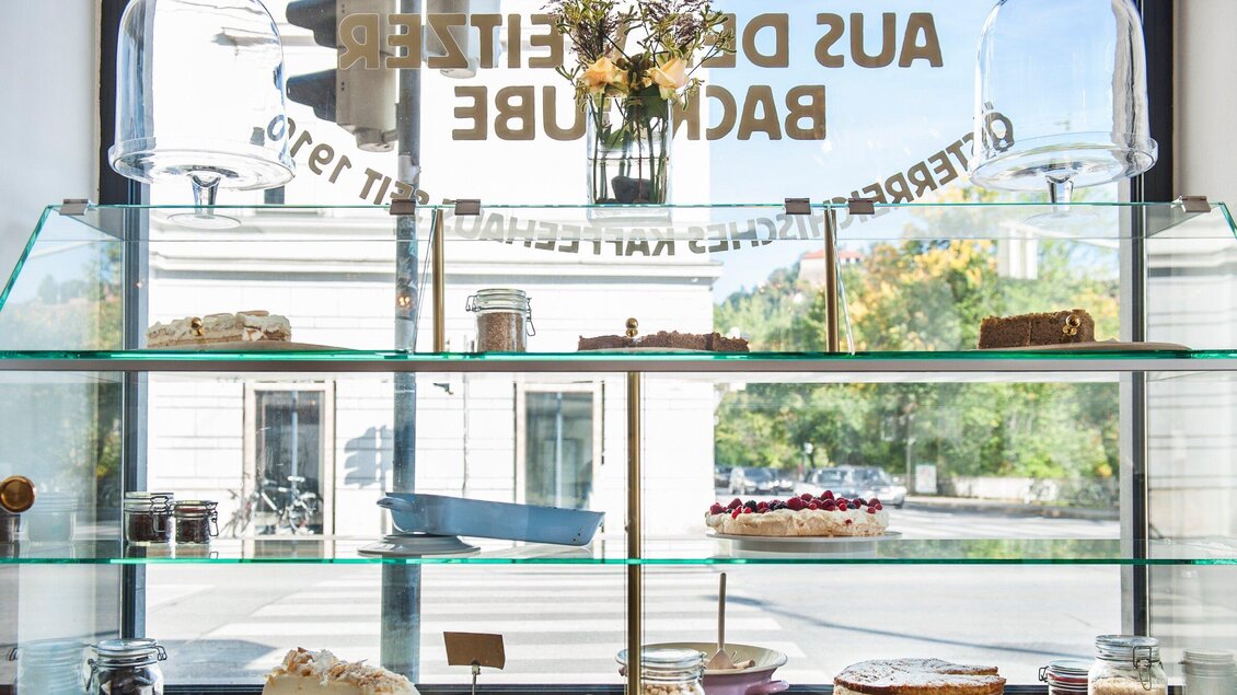 Eine Vitrine mit verschiedenen Kuchen und Gebäck. Im Hintergrund sind blühende Pflanzen und eine helle, freundliche Atmosphäre zu sehen. | © Weitzer