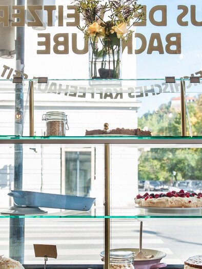 A display case with various cakes and pastries. In the background, blooming plants and a bright, friendly atmosphere can be seen. | © Weitzer
