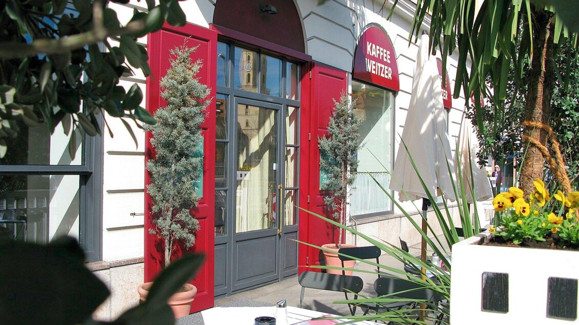 Ein einladendes Café mit roter Fassade und Pflanzen davor. Tische und Stühle stehen draußen für Gäste bereit. | © Weitzer