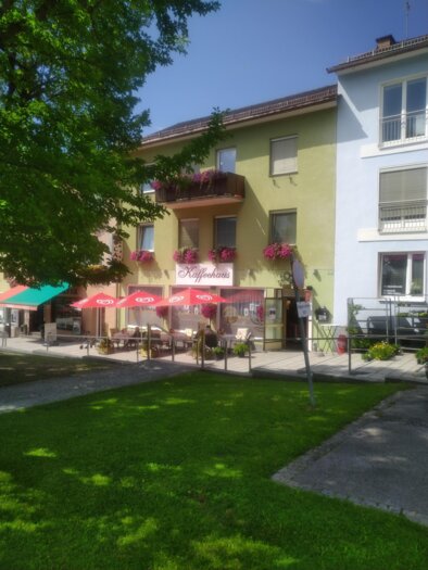 Ein charmantes Gebäude mit bunten Balkonen und Blumen. Vor dem Haus stehen Tische mit Sonnenschirmen im Freien. | © Peer Dieter