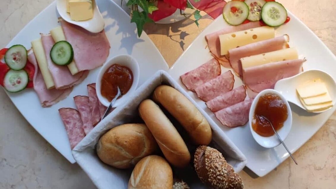 Ein Frühstücksbrett mit verschiedenen Wurst- und Käsesorten, frischen Gurkenscheiben und Brötchen. Dazu gibt es Marmelade in kleinen Schälchen. | © Peer Dieter