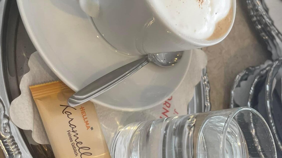 Eine Tasse Kaffee mit Schaum und einer Prise Kakao, serviert auf einem silbernen Tablett. Dazu ein Glas Wasser und ein kleines Plätzchen. | © Peer Dieter