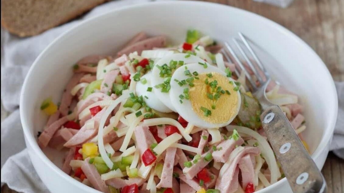 Ein farbenfrohes Nudelgericht mit Schinken, Paprika und einem weich gekochten Ei. Daneben liegt frisches Brot auf einem Holzuntergrund. | © Peer Dieter
