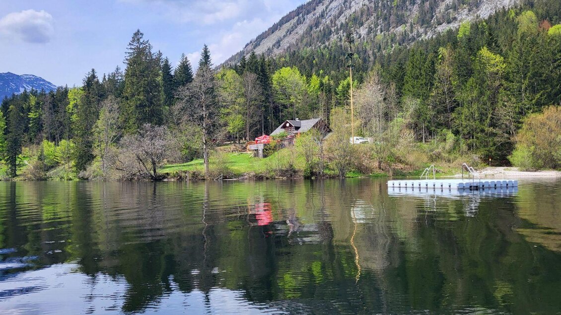 Eine ruhige Landschaft mit einem See und einer idyllischen Hütte umgeben von Bäumen. Im Hintergrund sind sanfte Berge und ein Schwimmsteg zu sehen. | © Petra Kirchschlager