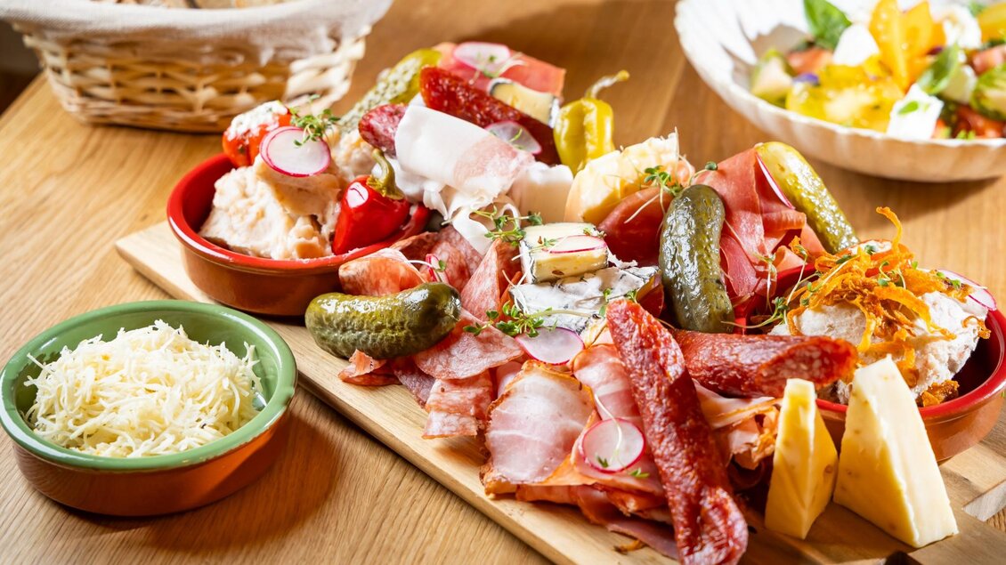 Holzbrett mit steirischem Aufschnitt, Käse, Aufstrichen, Essiggurkerln, Kren und Brotkorb im Hintergrund | © Kaiser Greißler - Werner Krug