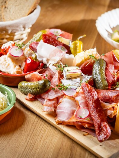 Holzbrett mit steirischem Aufschnitt, Käse, Aufstrichen, Essiggurkerln, Kren und Brotkorb im Hintergrund | © Kaiser Greißler - Werner Krug