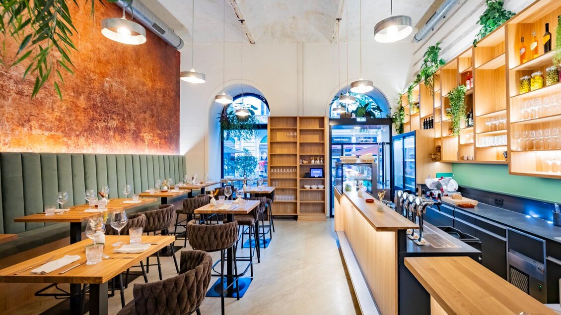 Innenraum eines hellen Lokals mit hoher Decke, Holztischen, Polsterbänken, Barbereich und Regalwand | © Kaiser Greißler - Werner Krug