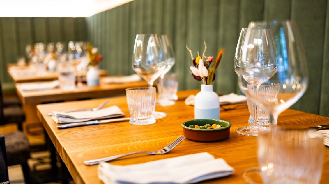 Klare Sicht auf gedeckte Holztische mit Trinkgläsern, Besteck und Stoffservietten vor gepolsterter Wand | © Kaiser Greißler - Werner Krug
