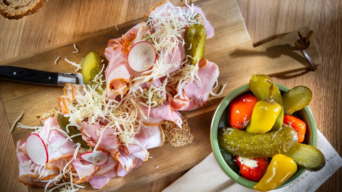 Holzbrett mit Brot, Schinken, Kren, Gurkerln, Senf und Radieschen, daneben Schüssel mit eingelegtem Gemüse | © Kaiser Greißler - Werner Krug