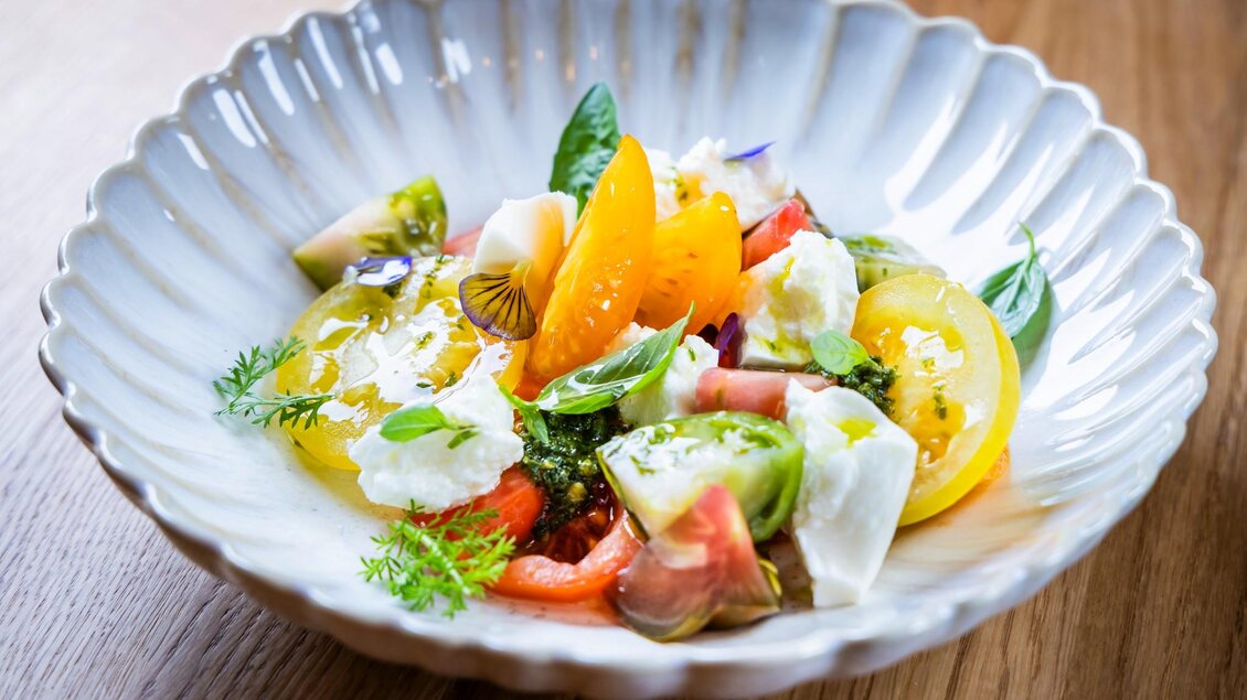Teller mit buntem Tomatensalat, Mozzarella, Kräutern und essbaren Blüten auf einem Holztisch | © Kaiser Greißler - Werner Krug