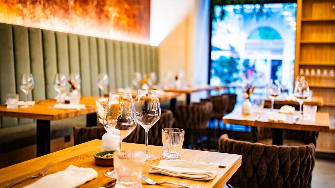 Nahaufnahme eines gedeckten Holztischs mit Gläsern, Besteck und Stoffserviette vor Polsterwand und Fenster | © Kaiser Greißler - Werner Krug