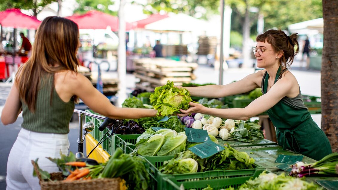 Eine Frau mit Fahrradkorb voller Gemüse kauft an einem Marktstand einen großen grünen Salat von einer Verkäuferin im grünen Schürze, während im Hintergrund weitere Stände mit frischem Gemüse zu sehen sind | © Graz Tourismus - Tom Lamm