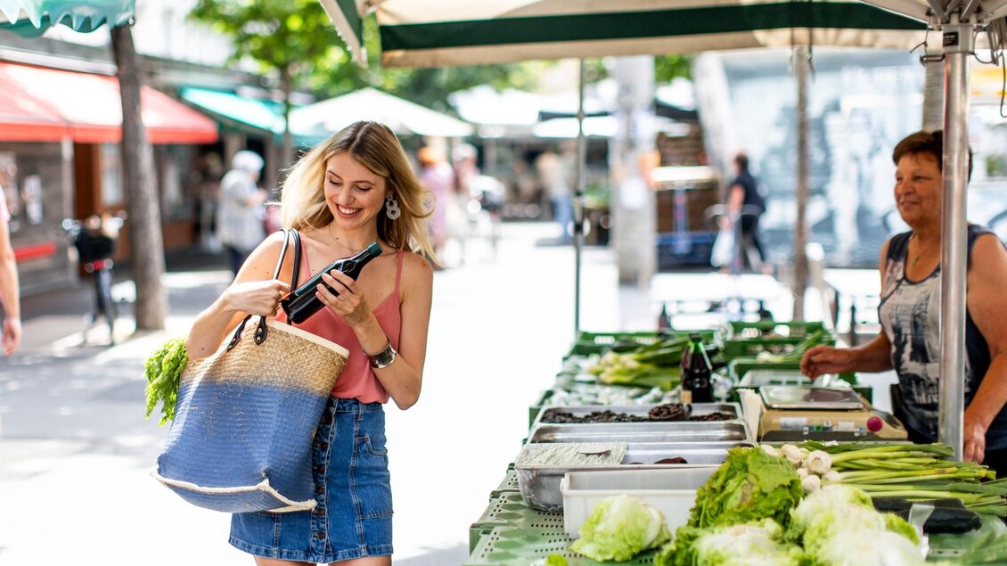 Frau mit Einkaufskorb hält eine Flasche Kürbiskernöl, vor einem Marktstand mit Salat, Zucchini und Frühlingszwiebeln | © Graz Tourismus - Tom Lamm