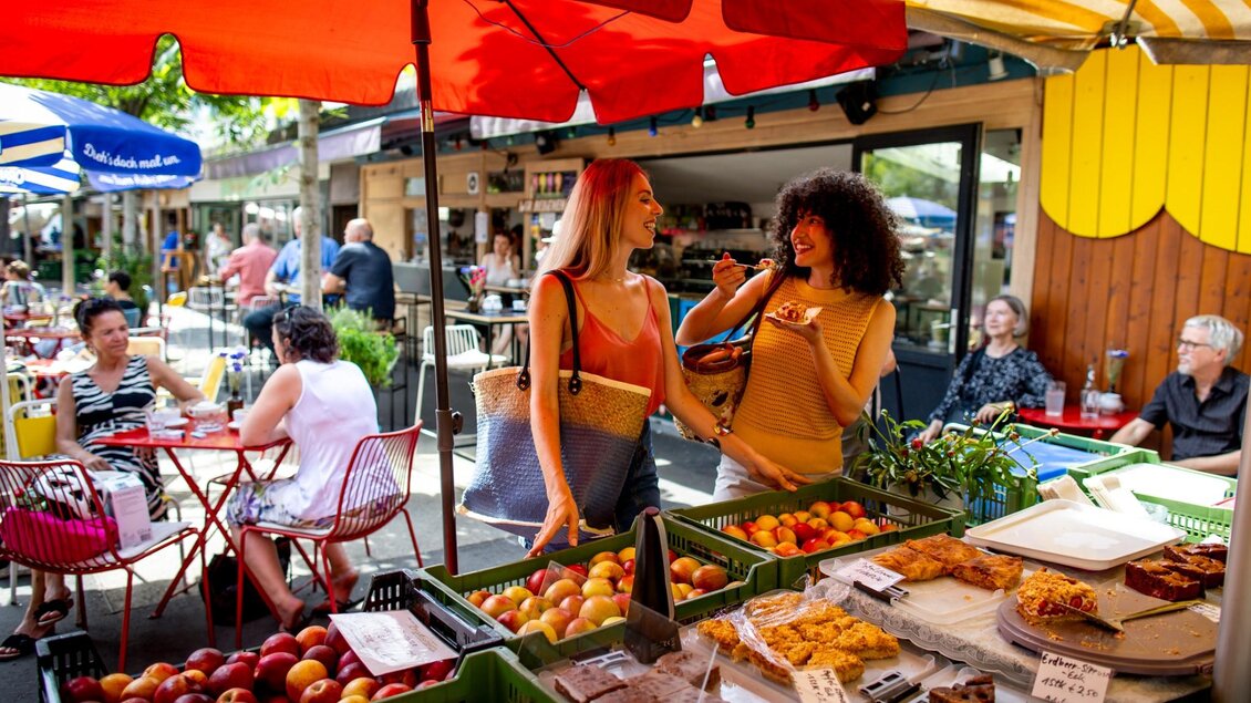 Zwei Frauen stehen an einem Marktstand mit Obst und Kuchen, eine probiert ein Stück Gebäck | © Graz Tourismus - Tom Lamm