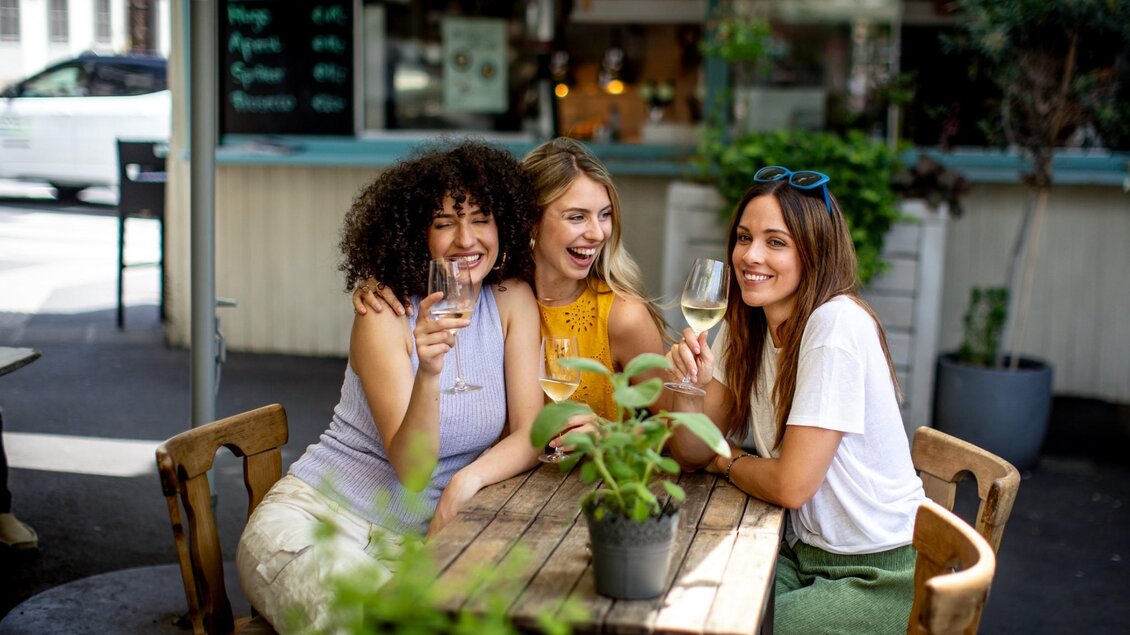 Drei junge Frauen sitzen lachend an einem Holztisch im Freien und stoßen mit Gläsern Weißwein an | © Graz Tourismus - Tom Lamm