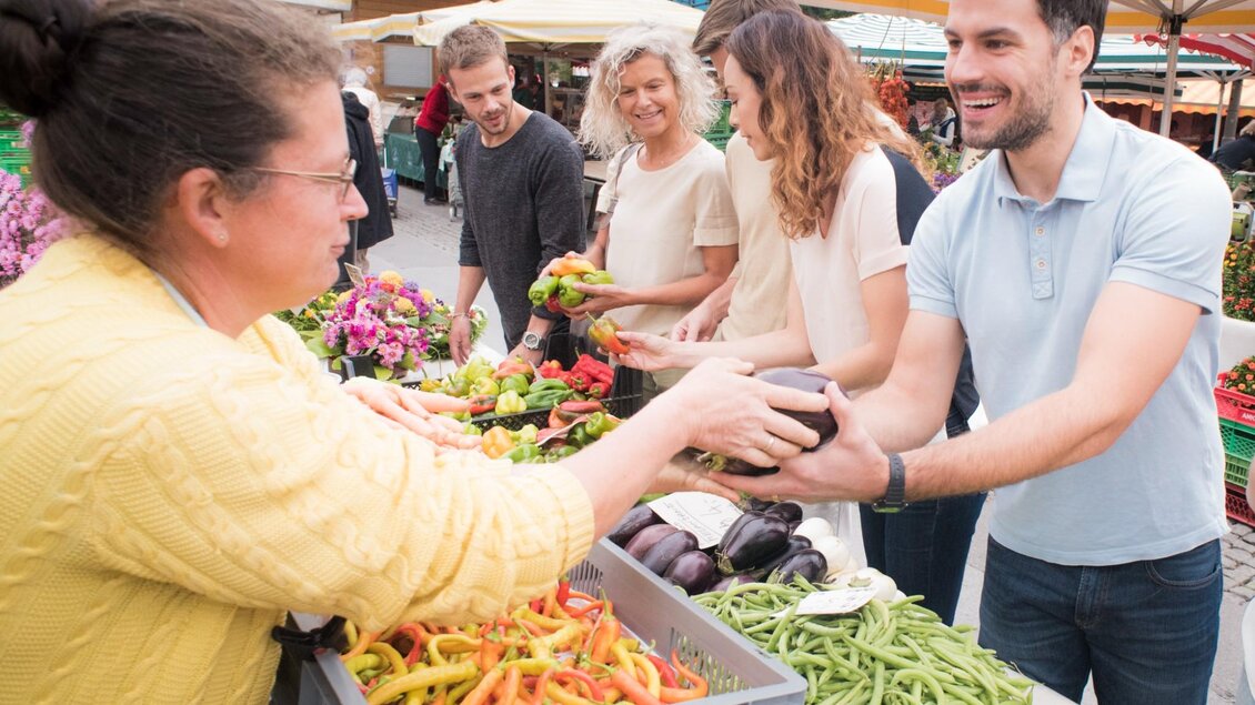 Verkäuferin reicht einem Mann eine Aubergine über den Marktstand mit Bohnen und Paprika. | © Graz Tourismus - Gerald Plattner