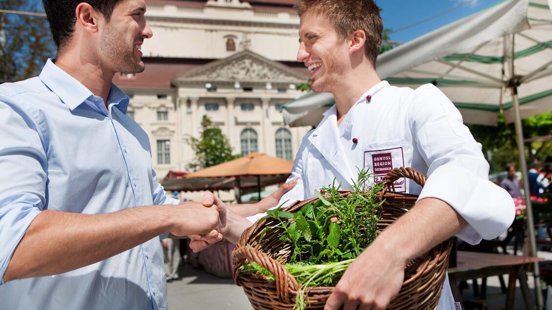 Koch mit Kräuterkorb begrüßt Besucher auf dem Kaiser-Josef-Markt in Graz, im Hintergrud ist die Grazer Oper erkennbar. | © Graz Tourismus - Harald Eisenberger