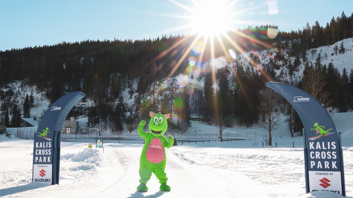 Ein grünes Maskottchen steht auf einem verschneiten Weg im Kalis Cross Park. Im Hintergrund strahlt die Sonne über den verschneiten Bergen. | © Loretta Kvitek