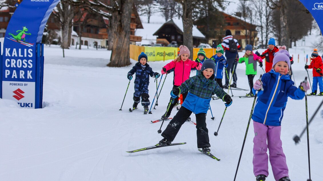 Eine Gruppe von Kindern fährt Ski im Schnee. Im Hintergrund sind Bäume und Gebäude zu sehen. | © Loretta Kvitek