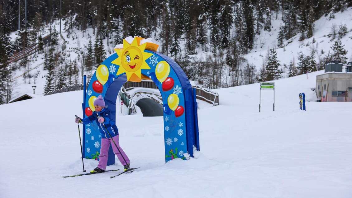 Ein Kind in blauer und pinker Skiausrüstung fährt unter einem bunten Tor mit einer Sonne hindurch. Die Umgebung ist schneebedeckt und von Tannenwäldern umgeben. | © Loretta Kvitek