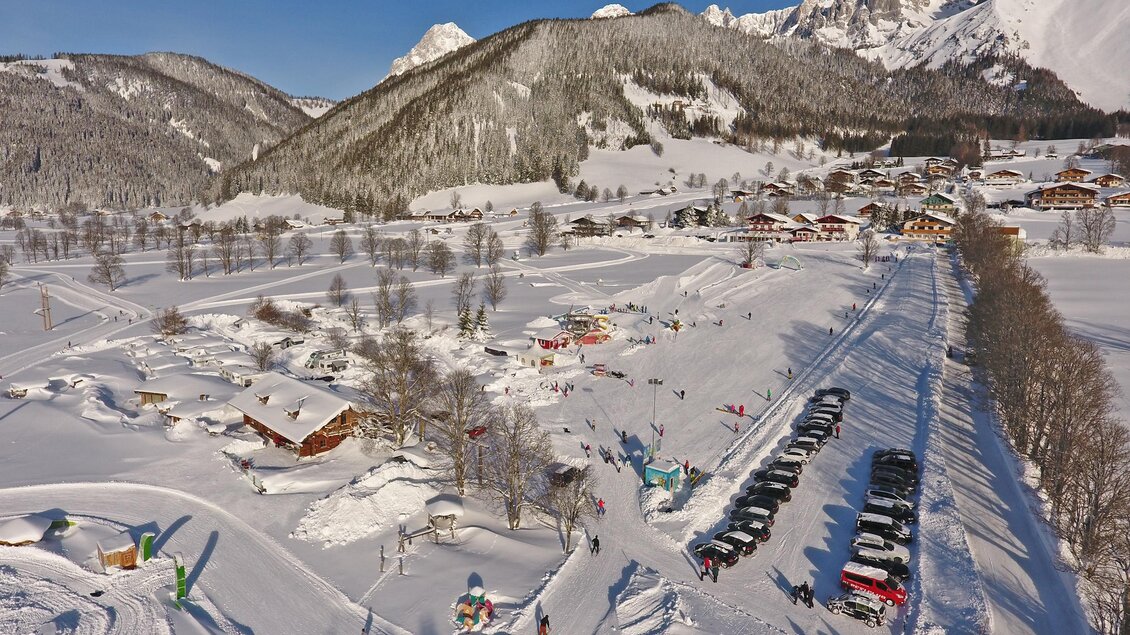 Eine Winterlandschaft mit Schnee bedeckten Bergen und vielen Skifahrern. Im Vordergrund sind Hütten und geparkte Autos entlang einer verschneiten Straße. | © Hans-Peter Steiner