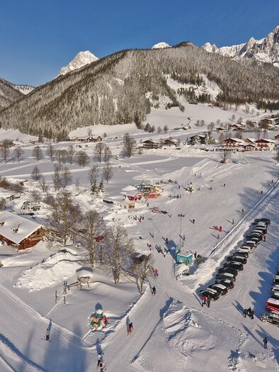 Eine Winterlandschaft mit Schnee bedeckten Bergen und vielen Skifahrern. Im Vordergrund sind Hütten und geparkte Autos entlang einer verschneiten Straße. | © Hans-Peter Steiner