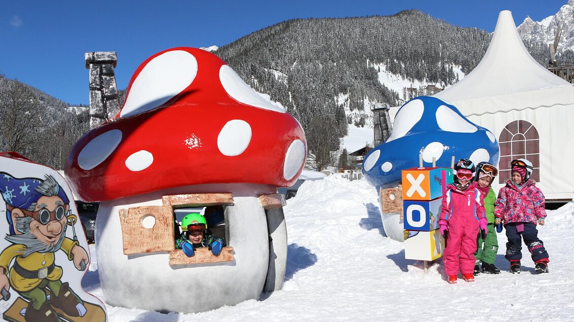 Bunte Pilzhäuser im Schnee mit fröhlichen Kindern. Im Hintergrund sind verschneite Berge und ein Zelt zu sehen. | © Hans-Peter Steiner