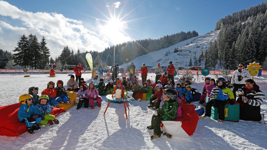 Eine Gruppe von Kindern sitzt im Schnee und genießt die Wintersonne. Im Hintergrund sind verschneite Berge und einige Betreuer zu sehen. | © Hans-Peter Steiner