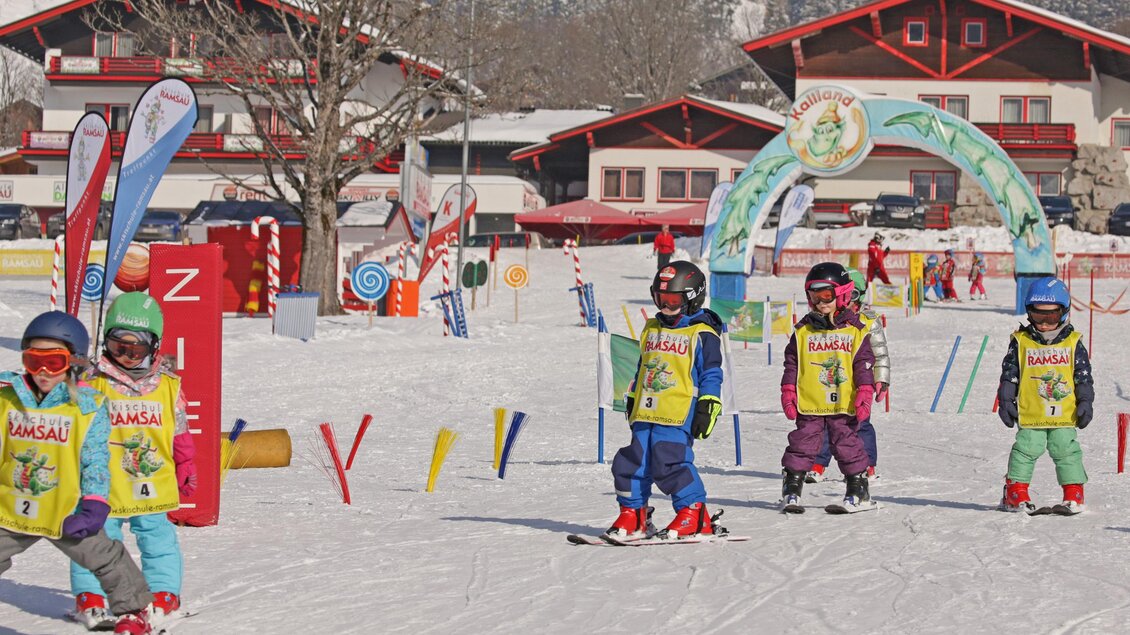 Eine Gruppe von Kindern in bunten Skianzügen lernt Skifahren auf einer verschneiten Piste. Im Hintergrund sind Holzhäuser und bunte Fahnen zu sehen. | © Hans-Peter Steiner