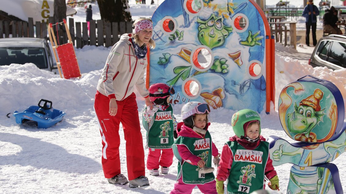 Eine lustige Schneespielplatz mit Kindern, die Skiausrüstung tragen. Im Hintergrund ist eine bunte Spielstation mit einem Monster-Motiv zu sehen. | © Hans-Peter Steiner