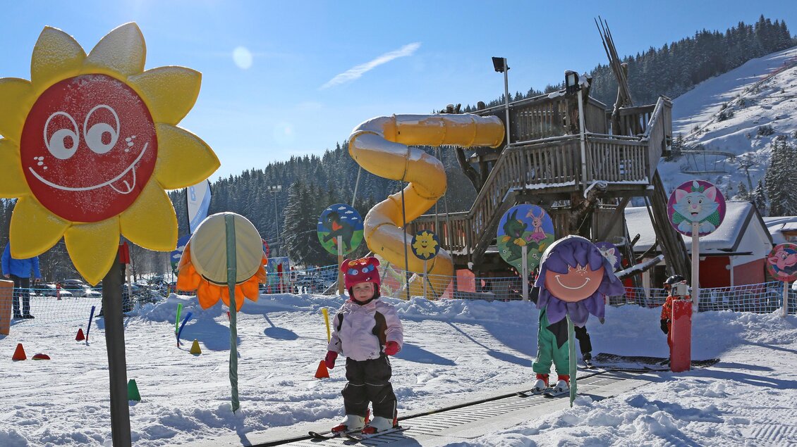 Ein winterlicher Spielplatz mit bunten Schildern und einer Rutsche. Kinder spielen im Schnee unter klarem Himmel. | © Hans-Peter Steiner