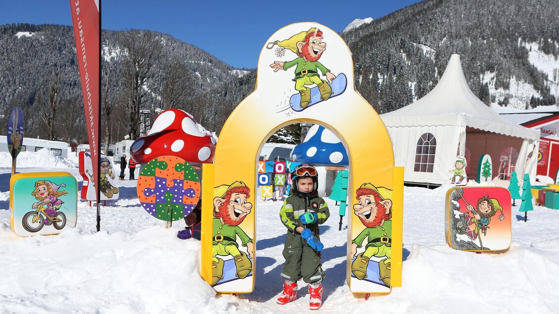 Ein Kind steht vor einem bunten Spielbogen im Schnee. Im Hintergrund sind fröhliche Figuren und eine Zeltstruktur zu sehen. | © Hans-Peter Steiner