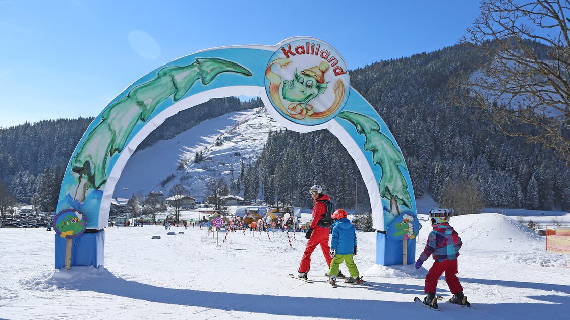 Ein bunter Torbogen mit der Aufschrift "Kalli Land" steht vor einem schneebedeckten Berg. Kinder in Skikleidung gehen fröhlich in Richtung des Skigebiets. | © Hans-Peter Steiner