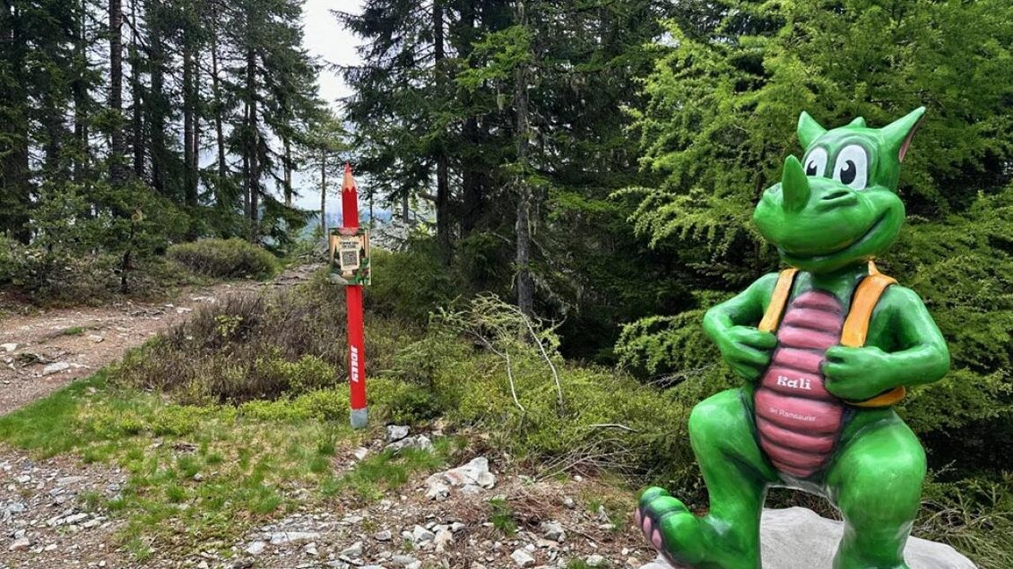 Eine grüne Drachenfigur steht auf einem Stein im Wald. Im Hintergrund ist ein Wanderweg mit einer roten Markierung sichtbar. | © Rittisberg