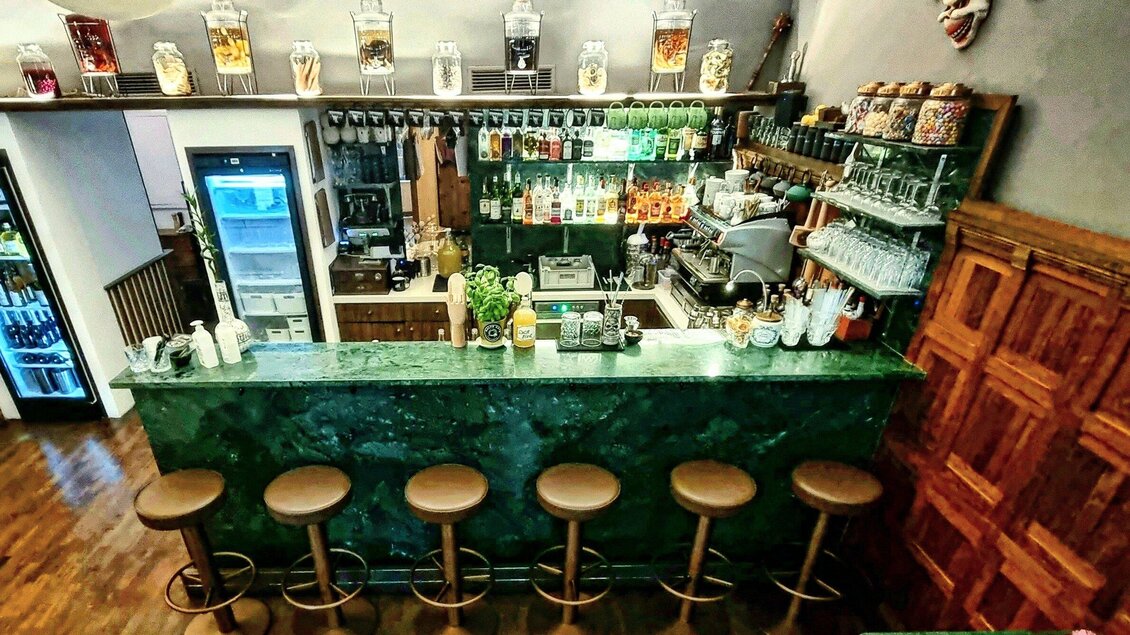 Eine stilvolle Bar mit einer grünen Theke und hölzernen Barhockern. Im Hintergrund sind zahlreiche Spirituosen und Gläser auf einem Regal angeordnet. | © Kalte Ente