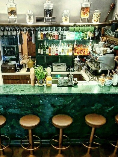 A stylish bar with a green counter and wooden bar stools. In the background, numerous spirits and glasses are arranged on a shelf. | © Kalte Ente