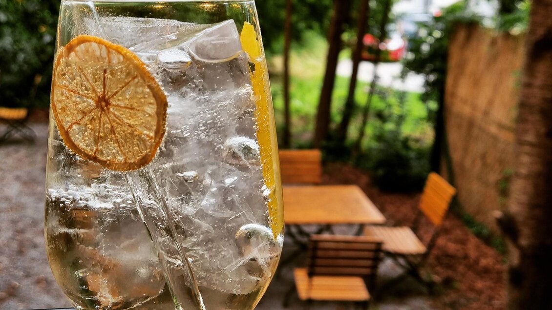 Ein erfrischendes Getränk in einem hohen Glas mit Eis und einer Scheibe Zitrone. Im Hintergrund sind Bäume und gemütliche Sitzgelegenheiten zu sehen. | © Kalte Ente