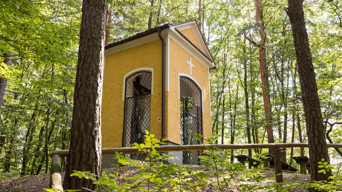 Ein kleines, gelbes Bauwerk im Wald mit Fenstern und einem Kreuz an der Vorderseite. Umgeben von hohen Bäumen und grünen Pflanzen. | © TV Region Graz/Harry Schiffer