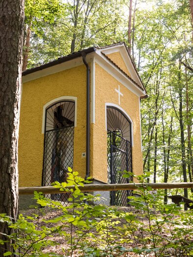 Ein kleines, gelbes Bauwerk im Wald mit Fenstern und einem Kreuz an der Vorderseite. Umgeben von hohen Bäumen und grünen Pflanzen. | © TV Region Graz/Harry Schiffer