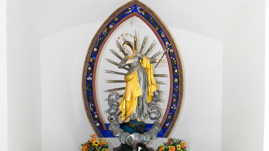 Eine Statue einer Heiligen in einer Nische, umgeben von bunten Blumen. Die Statue hat goldene und blaue Akzente und strahlt eine ruhige Atmosphäre aus. | © TV Region Graz/Harry Schiffer