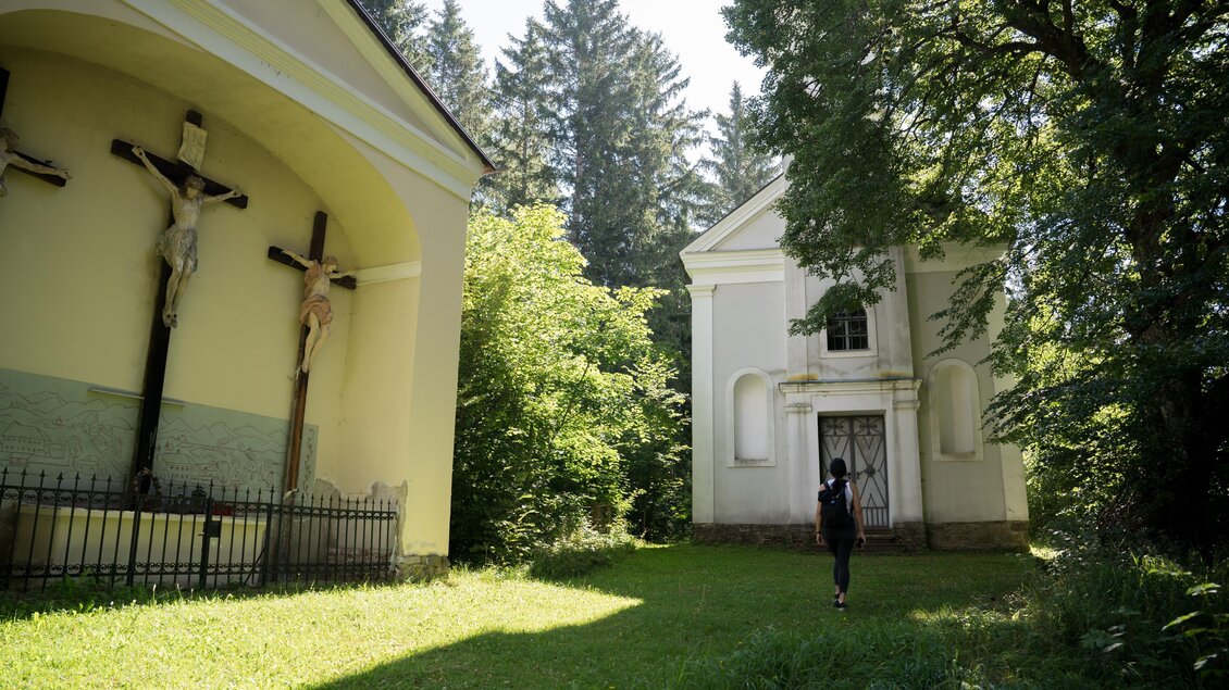 Eine ruhige Szene mit zwei alten Gebäuden inmitten von Bäumen. Im Vordergrund steht eine Person auf der grünen Wiese. | © Anita Fössl