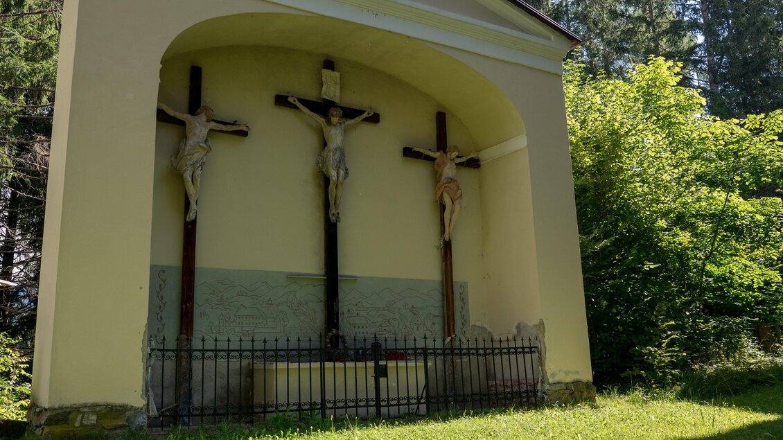 Eine kleine Kapelle mit drei Kreuzigungen steht unter einem Bogen. Die Umgebung ist grün und von Bäumen umgeben. | © Anita Fössl