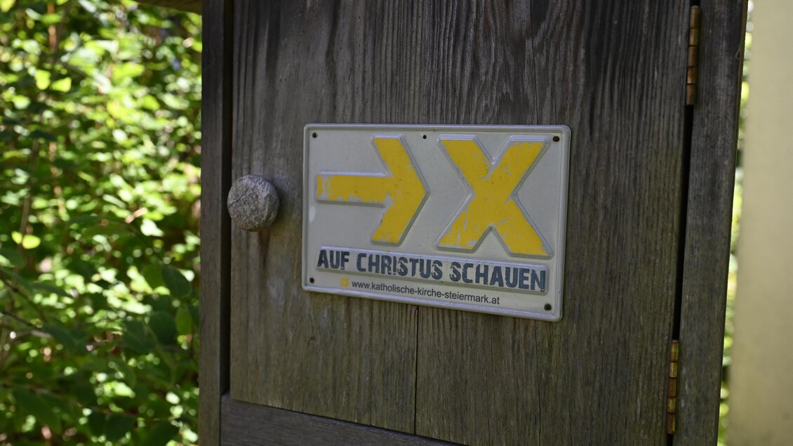 Ein Schild an einer Holzkonstruktion mit dem Aufdruck "AUF CHRISTUS SCHAUEN". Das Symbol zeigt nach rechts und ist in gelber Farbe gehalten. | © Anita Fössl