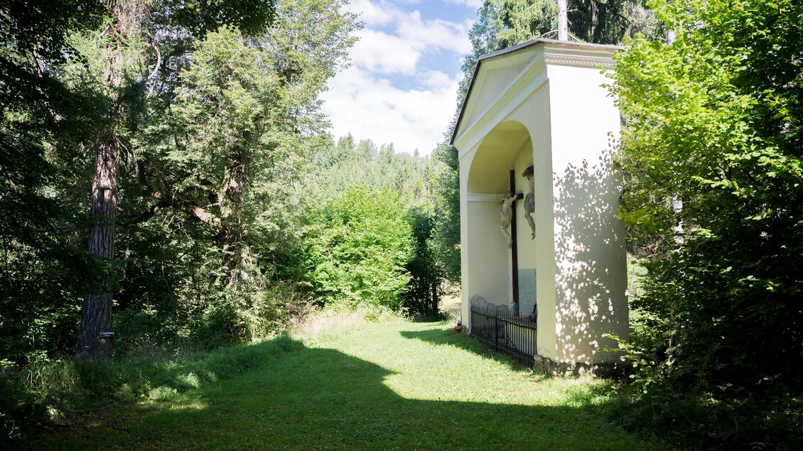Ein ruhiger Weg im Wald mit einer kleinen Kapelle. Umgeben von dichtem Grün und unter einem klaren Himmel. | © Anita Fössl