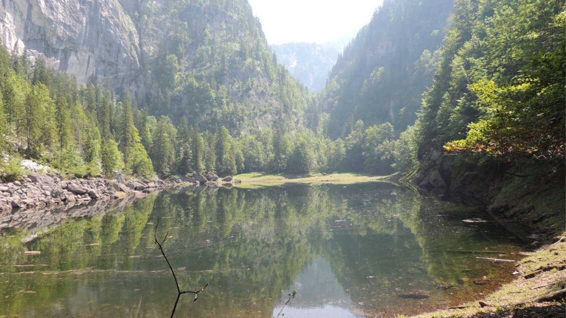Ein ruhiger See, umgeben von hohen Bergen und dichten Wäldern. Die Wasseroberfläche spiegelt die beeindruckende Landschaft wider.