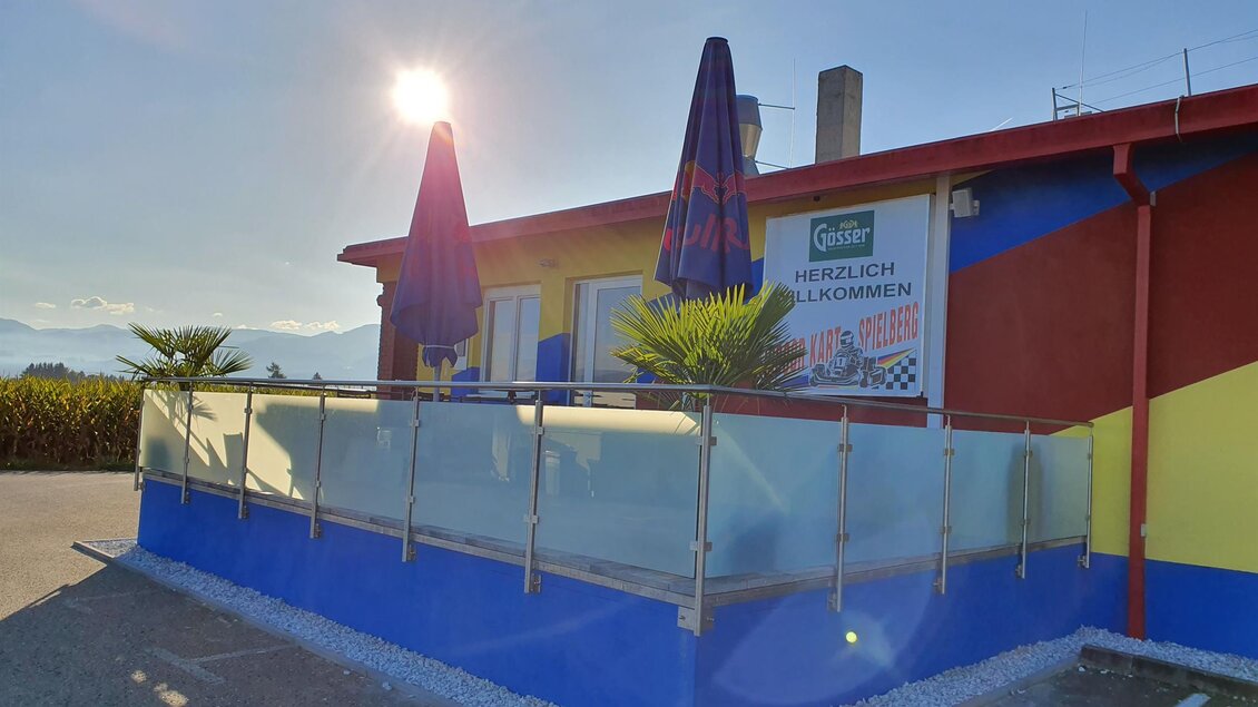 Ein farbenfrohes Gebäude mit einer Terrasse und Sonnenschirmen. Es gibt Palmen und eine klare Aussicht auf die Umgebung. | © Indoorkart Spielberg