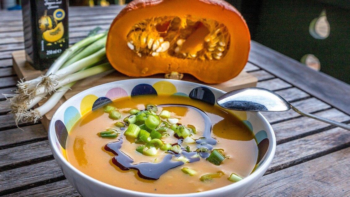 Eine köstliche Kürbissuppe in einer bunten Schüssel, garniert mit Frühlingszwiebeln. Im Hintergrund liegt ein halbierter Kürbis und ein Flasche Öl. | © Pixabay
