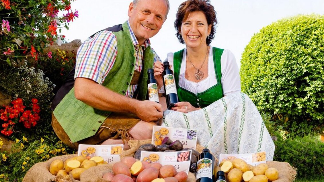 Ein Paar in traditioneller Tracht sitzt im Garten und hält Flaschen mit Bauernhofprodukten in der Hand. Um sie herum liegen verschiedene Kartoffelsorten und Präsentkörbe. | © Starzinger