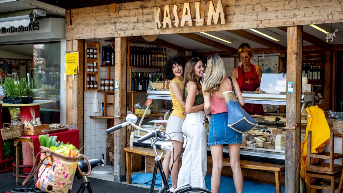 Drei Frauen kaufen an einem Marktstand mit Käse und Feinkost ein, Fahrrad mit Korb im Vordergrund | © Graz Tourismus - Tom Lamm